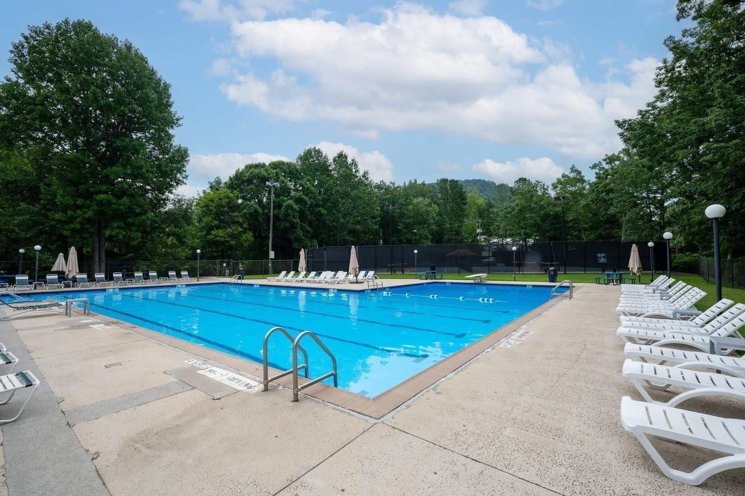 A rectangular swimming pool with clear blue water, surrounded by white lounge chairs and umbrellas, with trees and a grassy area in the background.