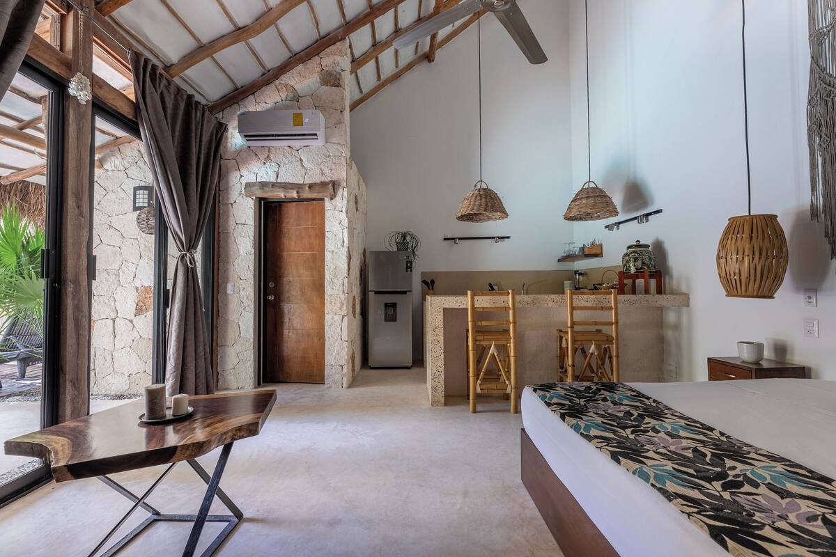 A bedroom with a bed, a desk, and a kitchenette in the background.