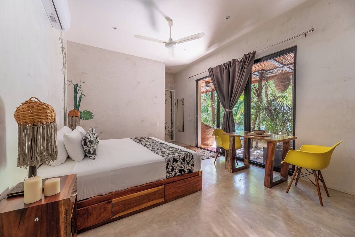 A modern bedroom with a bed, bedside table, and a dining area with chairs.