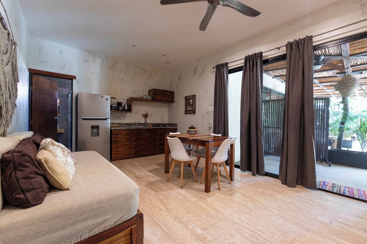 A compact living space with a kitchenette, a dining area with a table and chairs, a sofa with cushions, and a sliding glass door leading to an outdoor area.