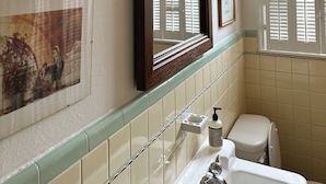 Ein Badezimmer mit Toilette, Waschbecken und Spiegel. Ein Fenster mit Rollläden, eine Wandleuchte und ein dekorativer Teller mit floralem Dekor.