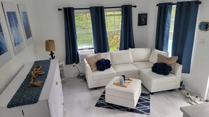 Una sala de estar moderna con un sofá blanco, una mesa de centro y una alfombra. Hay dos ventanas con cortinas azules, un espejo en la pared y una cita enmarcada.