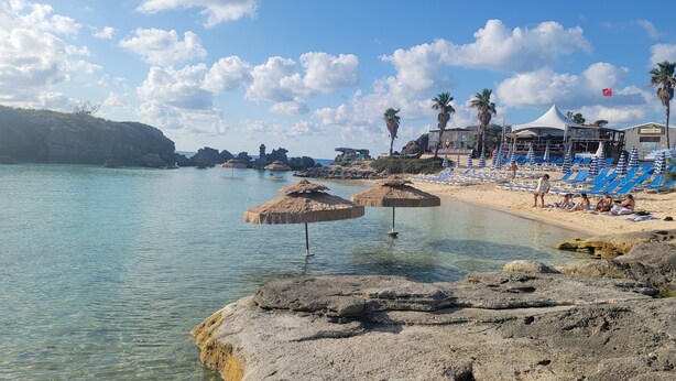 Una playa con agua cristalina, sombrillas de paja y sillas de descanso.