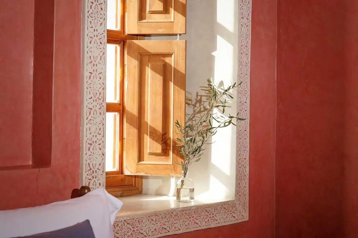 A room with a window, wooden shutters, and a vase with a plant.