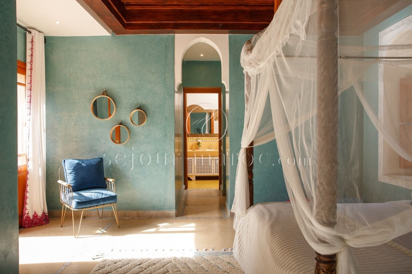 A bedroom with a four-poster bed, a blue chair, and a vanity with a mirror.