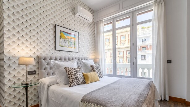 Ein Schlafzimmer mit einem gesteppten Kopfteil, einem großen Bett mit vielen Kissen, einem Nachttisch mit Lampe und einem großen Fenster mit Blick auf Gebäude.