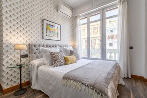 A bedroom with a tufted headboard, a large bed with multiple pillows, a bedside table with a lamp, and a large window with a view of buildings.
