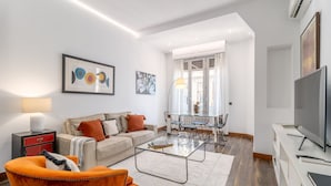 Ein modernes Wohnzimmer mit einer Couch, einem Couchtisch und einem orangefarbenen Sessel. Es gibt einen Essbereich mit einem Glastisch und Stühlen. Ein Fernseher ist an der Wand befestigt.