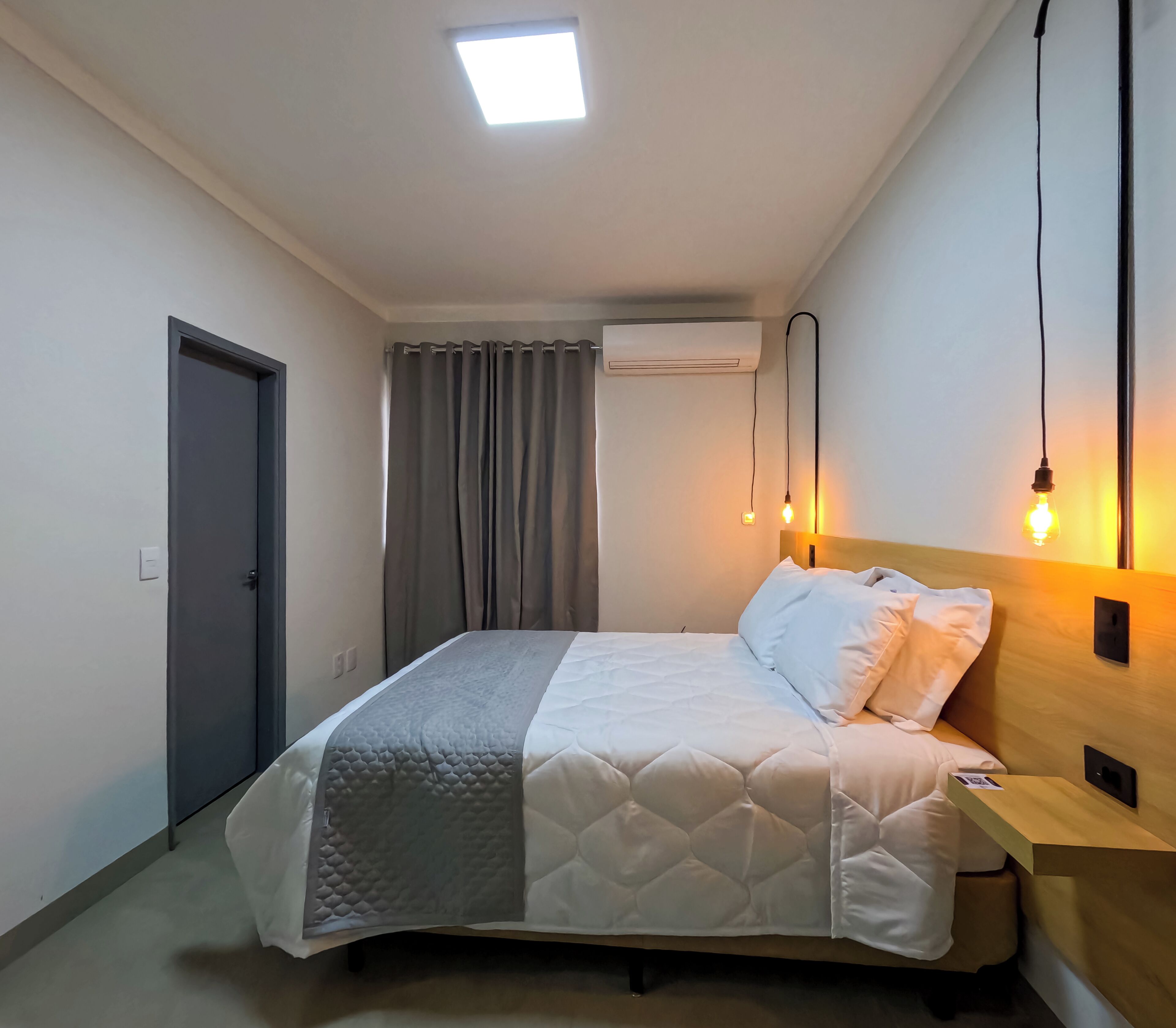 Ein ordentlich bezogenes Bett in einem Zimmer mit einer Tür, einer Klimaanlage und einem Nachttisch mit Lampe.