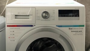A front-loading washing machine with a control panel, a power outlet, and a window above it.