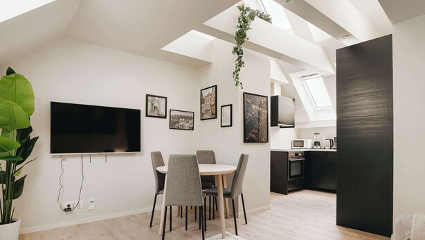A modern living room with a dining area, a flat-screen TV mounted on the wall, and a kitchen in the background.