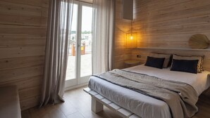 A bedroom with a wooden floor, a bed with a white and gray bedspread, a bench, a wall-mounted lamp, and a view of buildings through a glass door.