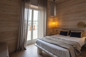 A bedroom with a wooden floor, a bed with a white and gray bedspread, a bench, a wall-mounted lamp, and a view of buildings through a glass door.