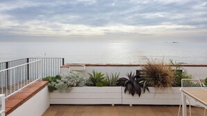 A terrace with a view of the sea, a white planter with various plants, a white table, and a white chair.
