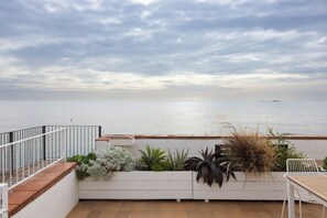 A terrace with a view of the sea, a white planter with various plants, a white table, and a white chair.