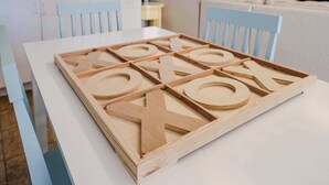 Ein Holz-Tic-Tac-Toe-Spiel auf einem weißen Tisch in einem Raum mit hellblauen Stühlen und einem weißen Sofa.