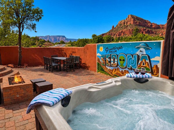 An outdoor hot tub with a mural, a brick fire pit, and a patio area with chairs and a table.
