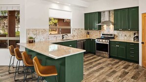 A modern kitchen with green cabinets, a large island with bar stools, a built-in oven, and a window overlooking a garden. - Myrinn| Yard w/ Hot Tub, Fire Pit & Red Rock Views (Sedona)