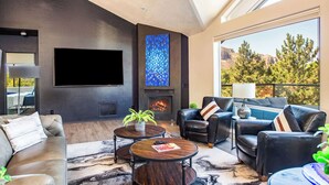 A modern living room with a fireplace, leather furniture, and a large window offering a scenic view. - Myrinn – Red Rock Views | Hot Tub, Gym & Billiards (Sedona)
