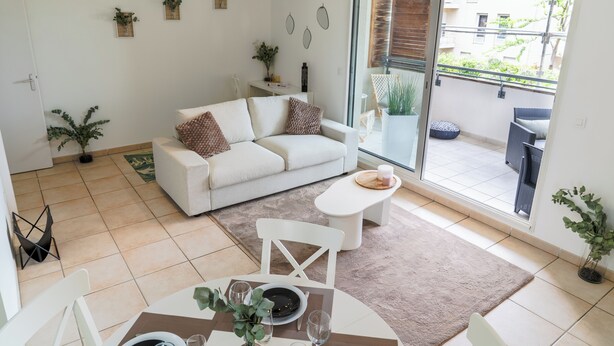 Ein modernes Wohnzimmer mit einem weißen Sofa, einem Esstisch mit Stühlen, einem Couchtisch und einer Glastür zum Balkon.
