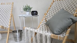 Zwei Makramee-Stühle mit Holzgestell und weißem Seilgeflecht, ein kleiner Tisch mit Topfpflanze, ein Weinglas und ein Eimer mit dem Aufdruck 'Champagner' auf einem Fliesenboden.
