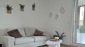 Ein modernes Wohnzimmer mit einem grauen Sofa, einem weißen Couchtisch und einem grauen Teppich. An den Wänden hängen drei Dekorationen und auf der Fensterbank steht ein Blumentopf.