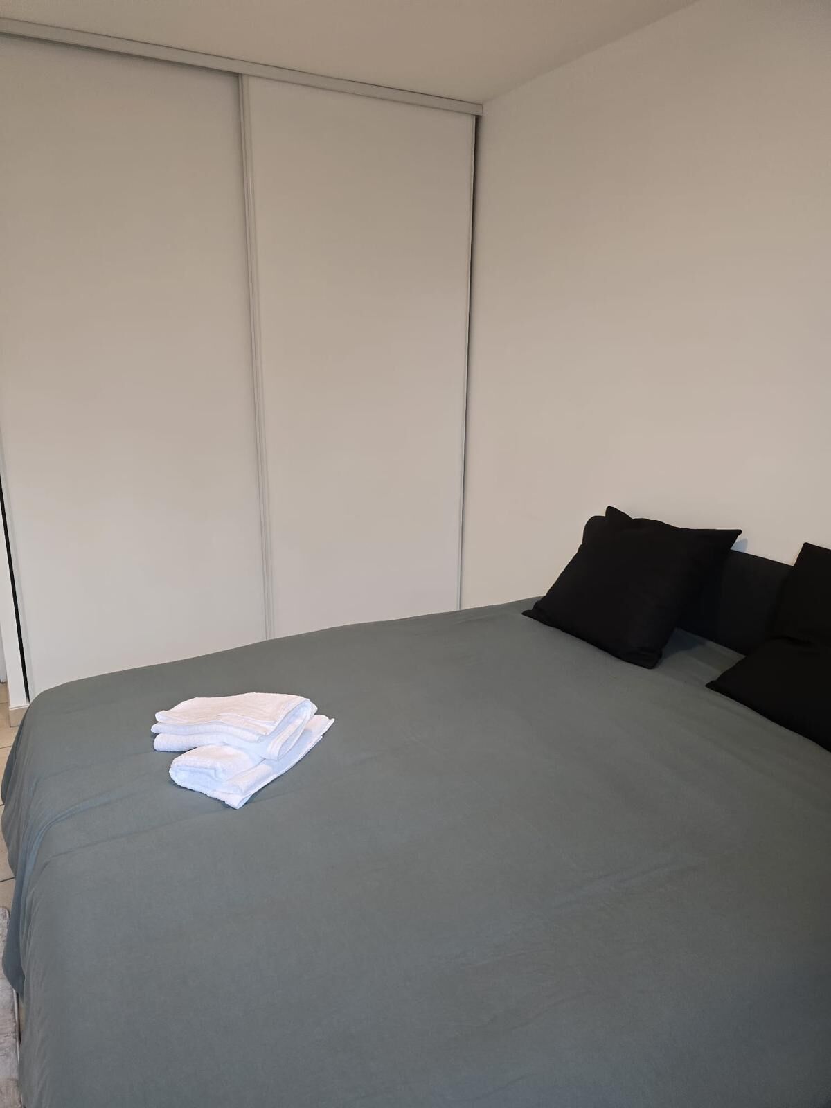 Ein ordentlich gemachtes Bett mit grauer Tagesdecke und einem aufgefalteten weißen Handtuch auf der linken Seite.