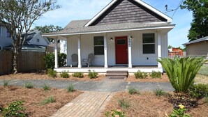 Une maison de plain-pied avec une porte d’entrée rouge, un porche couvert avec deux chaises et un chemin en briques menant à l’entrée.