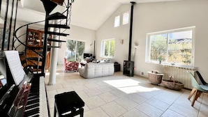 Un vaste salon agrémenté d’un piano, d’un escalier en colimaçon, d’une cheminée et de grandes fenêtres offrant une vue sur l’extérieur.