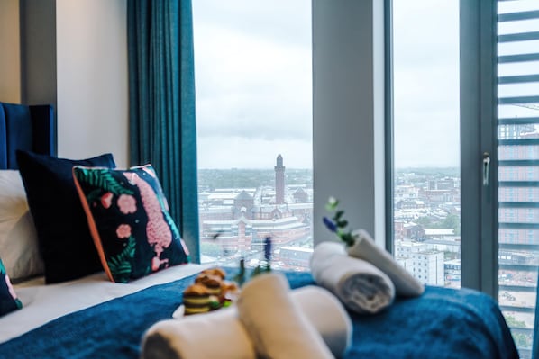 Une chambre d’hôtel dotée d’une grande fenêtre donnant sur la ville, d’un lit recouvert d’une couverture bleue et agrémentée de coussins décoratifs.