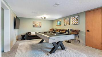 A pool table with a light wood finish and a pool cue, surrounded by four cushioned chairs with a brown leather finish.