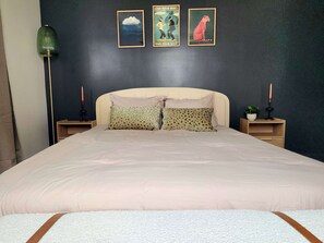 A bedroom with a large bed, two bedside tables, a floor lamp, and three framed posters on the wall.
