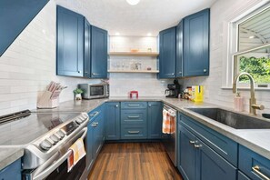 A modern kitchen with blue cabinets, a stainless steel stove, and a window with a view of greenery. - Fun Midcentury Home | Hot Tub | Karaoke | Games (Lancaster)