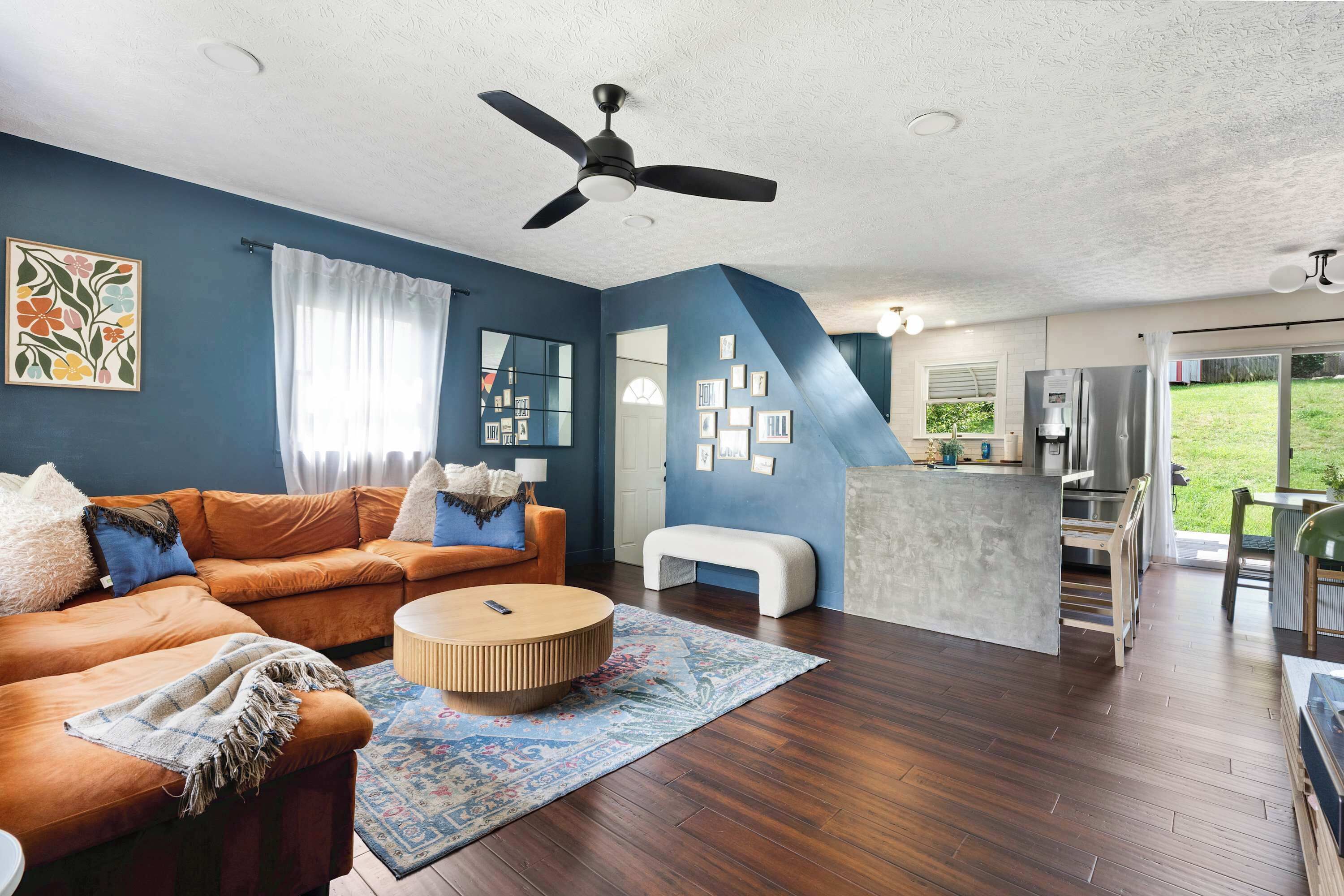 Un salon moderne avec un mur d’accent bleu, un ventilateur de plafond, un canapé, une table basse et un coin cuisine équipé d’un réfrigérateur et d’une table à manger.