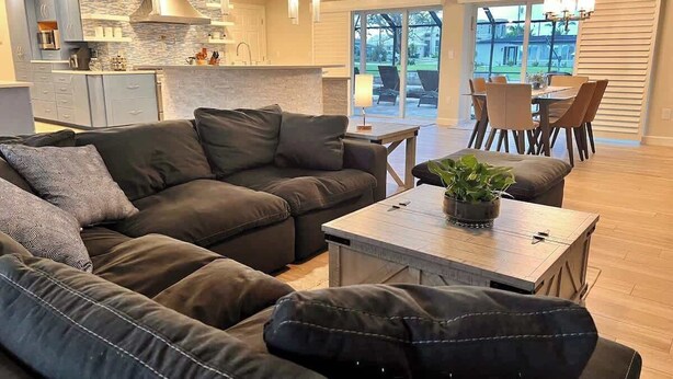 A modern living room with a sectional sofa, a coffee table with a plant, and a dining area with chairs and a chandelier.