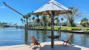 A waterfront deck with two wooden chairs and a small table, a gazebo, and a view of the water and surrounding buildings.