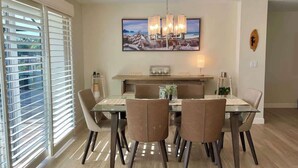 A dining area with a glass table, six chairs, a chandelier, and a painting above the table.