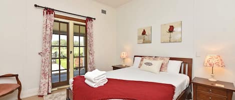 A bedroom with a bed, two bedside tables with lamps, a chair, a window with curtains, and two wall-mounted paintings.