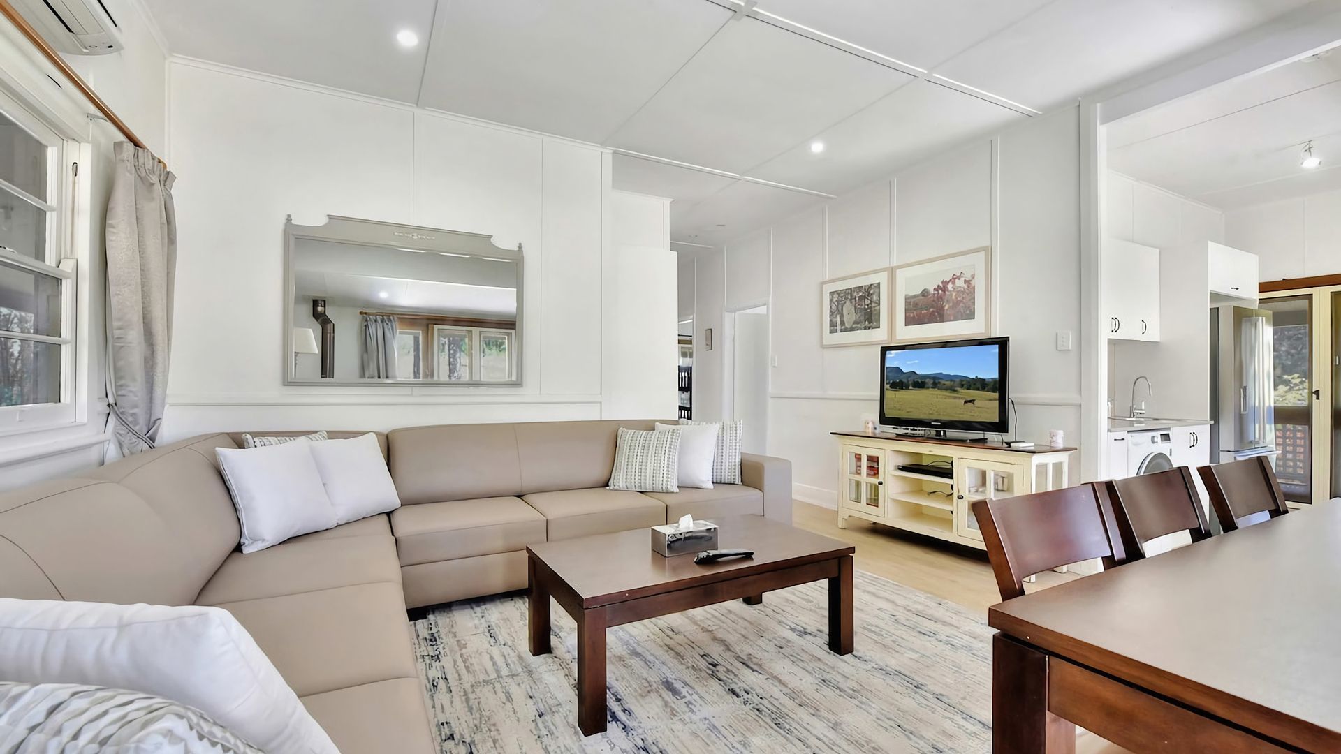 A modern living room with a sofa, coffee table, and TV. There is a dining area with chairs and a table. The room has white walls and a large mirror.