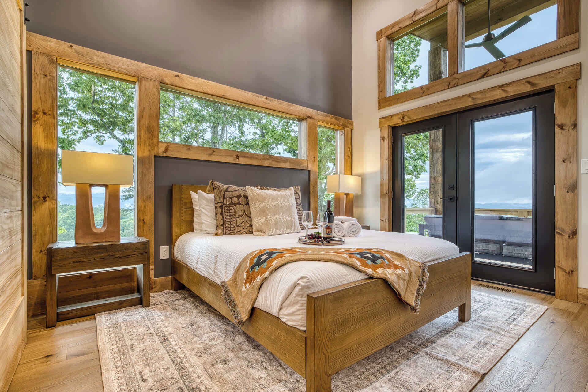 Une chambre à coucher avec un lit en bois, une table de chevet, une lampe et une vue sur l’extérieur grâce à de grandes fenêtres.