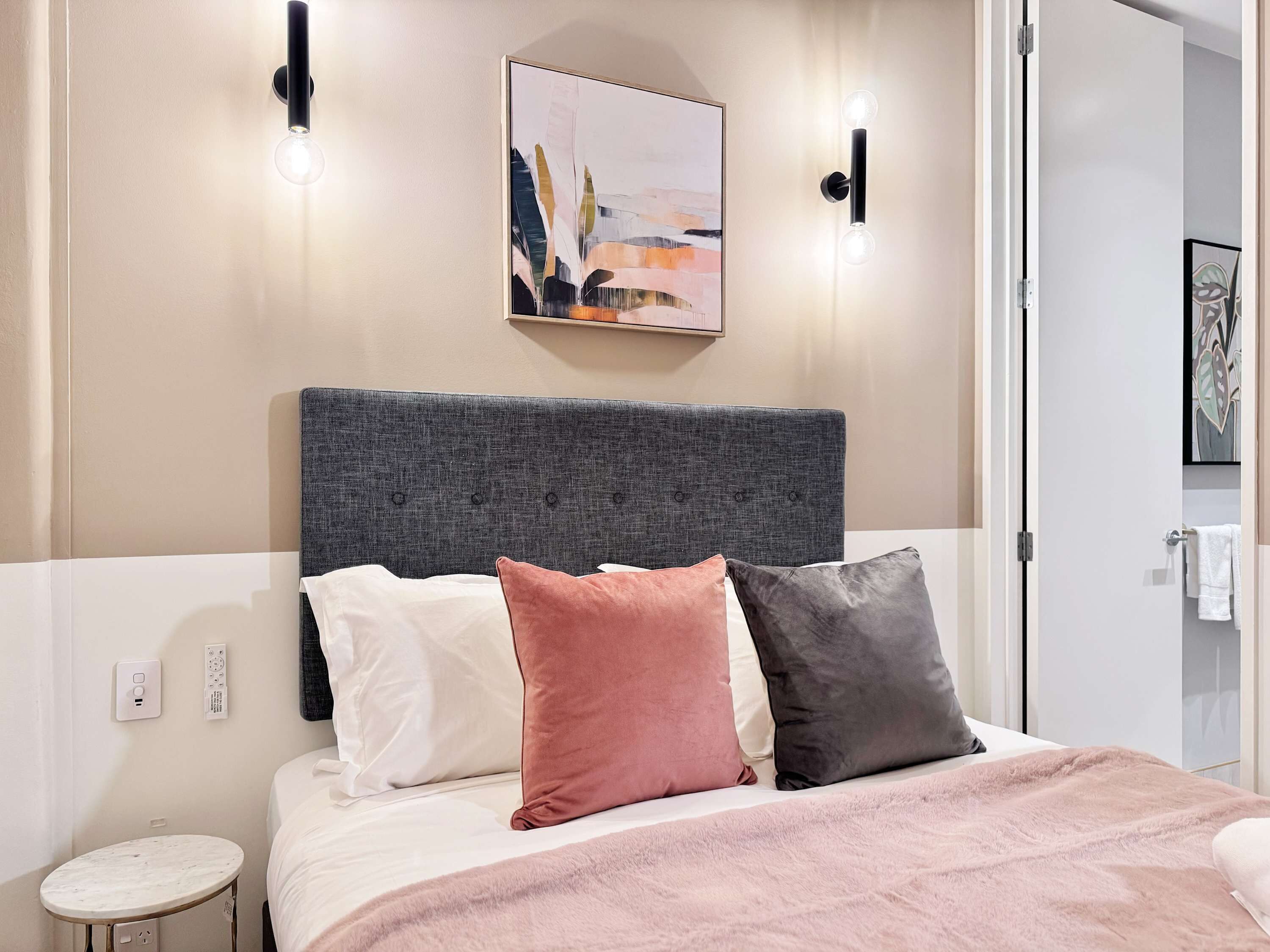 Une chambre à coucher moderne avec une tête de lit rembourrée de couleur grise, une couvre-lit blanche et trois coussins aux couleurs blanche, rose et grise. Deux appliques murales et un tableau abstrait encadré sont accrochés au mur au-dessus du lit.