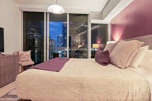 Quarto de hotel moderno com uma cama grande, uma mesinha de cabeceira, uma cadeira e vista para edifícios da cidade através de janelas do chão ao teto.