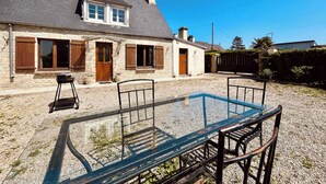 A two-story stone house with a gravel driveway and a glass table with metal chairs in the foreground. - La Maison d Utah , Beach and Memory (Sainte-Marie-du-Mont)