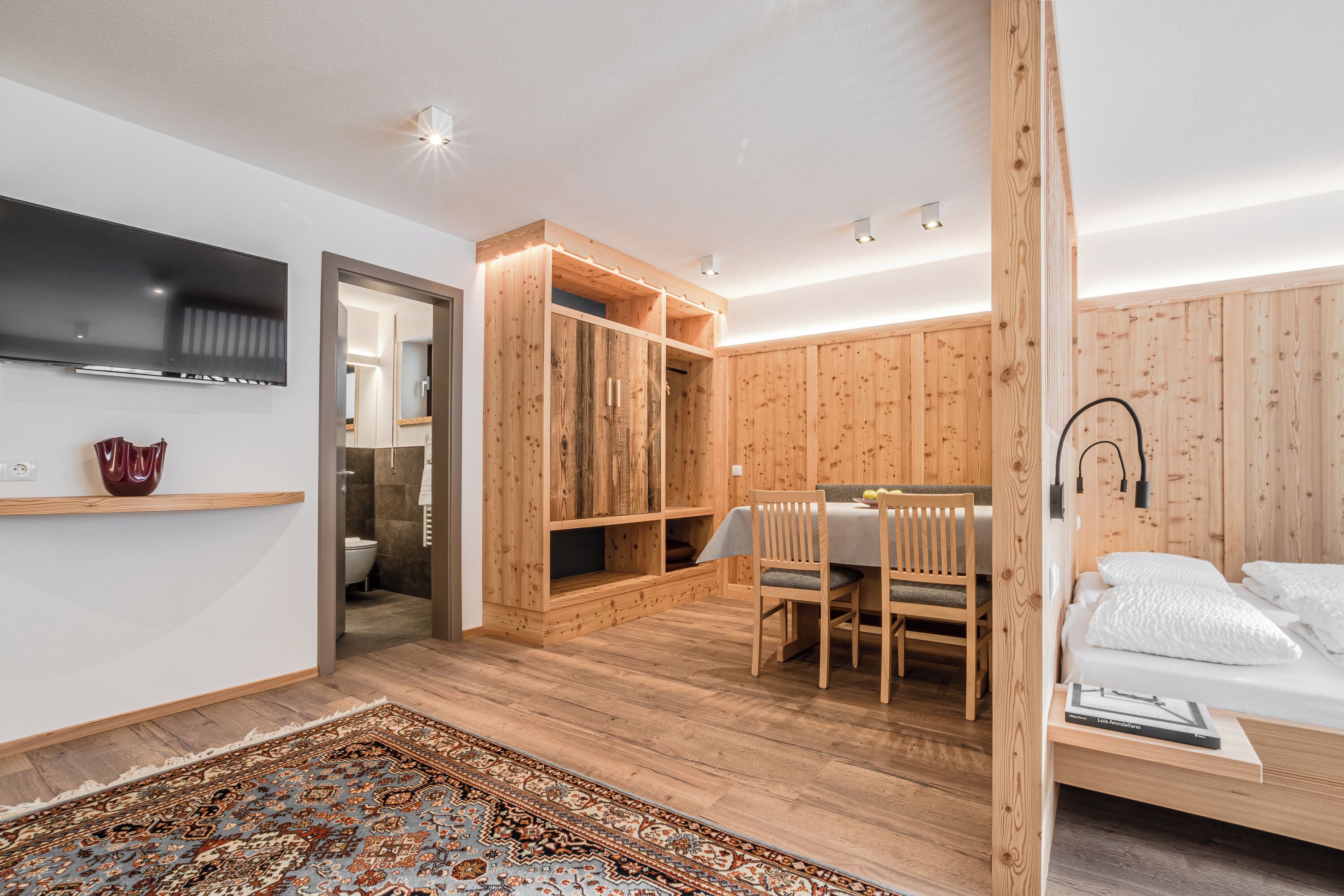 A modern bedroom with a wooden wardrobe, a dining area with a table and chairs, and a bed with a headboard.
