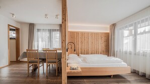 A bedroom with a wooden bed, a dining table with chairs, and a view of the outdoors through large windows. - Residence Ada (San Cassiano)