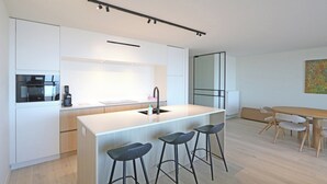 A modern kitchen with a central island, bar stools, a dining table with chairs, and a large window.