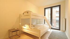 A single-bed room with a bunk bed, a bedside table, a lamp, a small stool, and a view of the outside balcony.