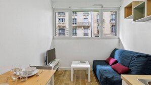 A compact living space with a dining area, a sofa, a TV, and a window with a view of buildings. - Well-equipped studio near the Parc des Princes (Paris)