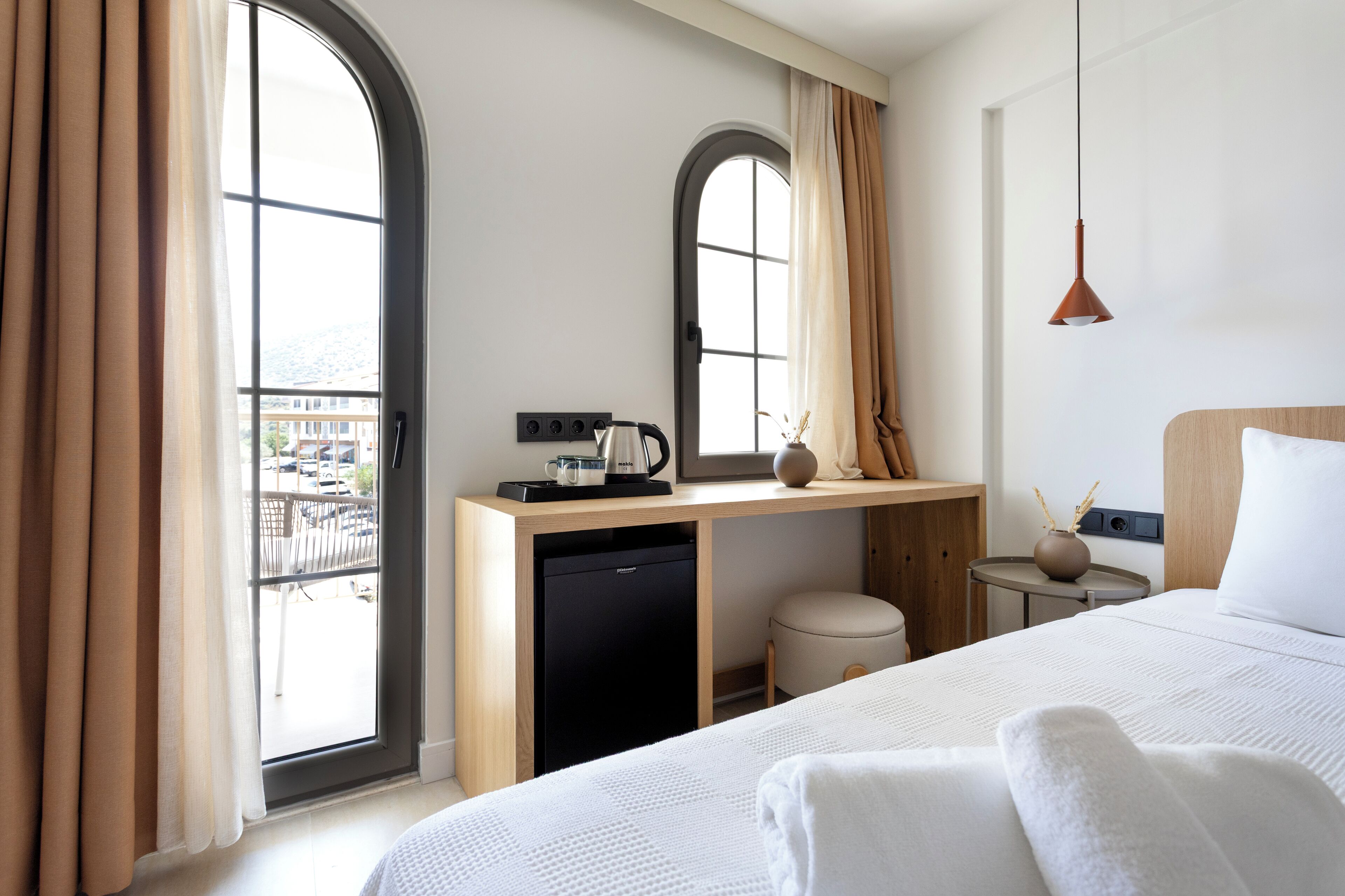 A hotel room with a bed, a desk with a coffee maker, a small stool, and a view of the outside.