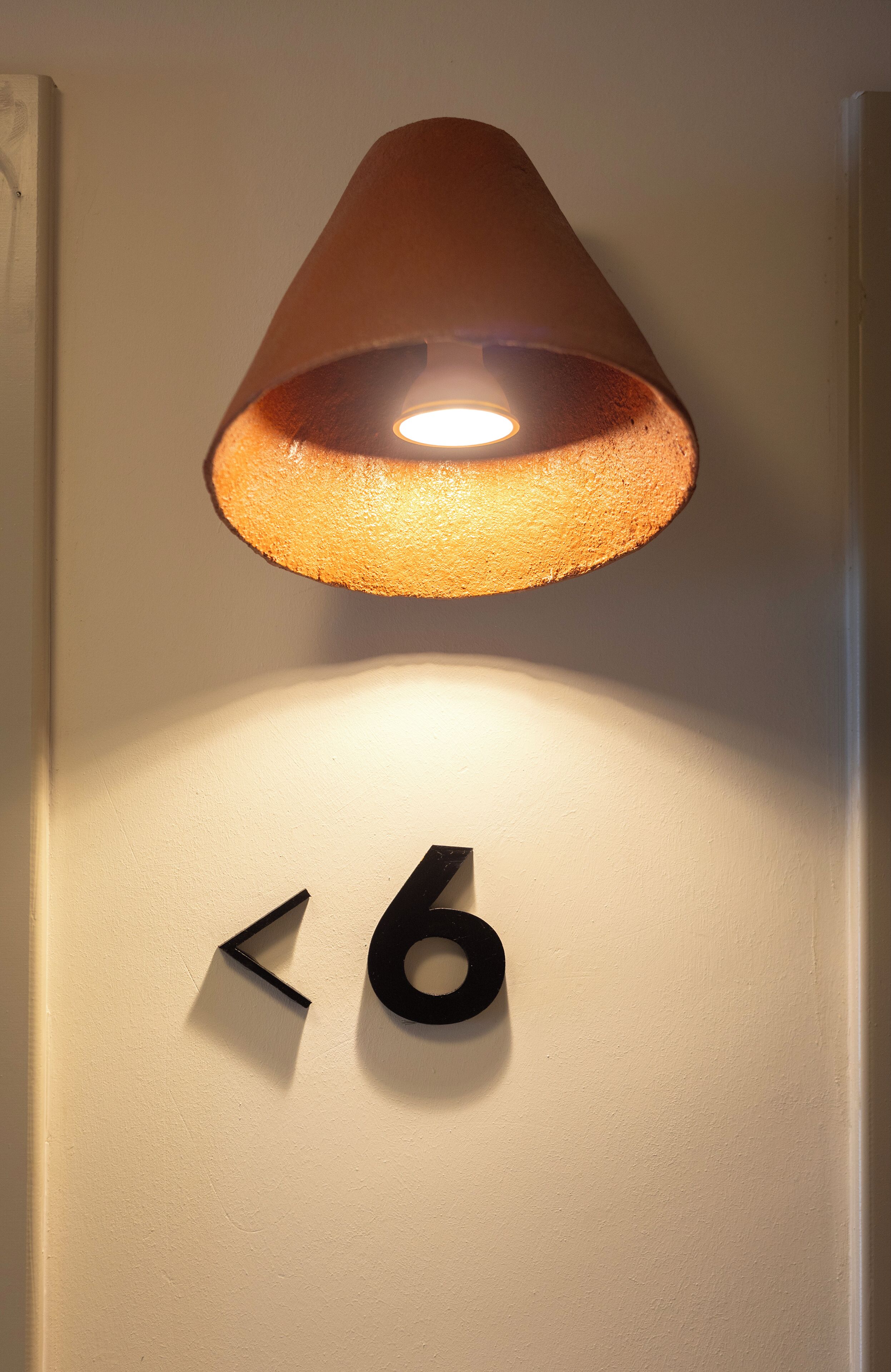 Uma luminária de parede com cúpula cônica iluminando uma parede com o número '6' e um símbolo triangular.